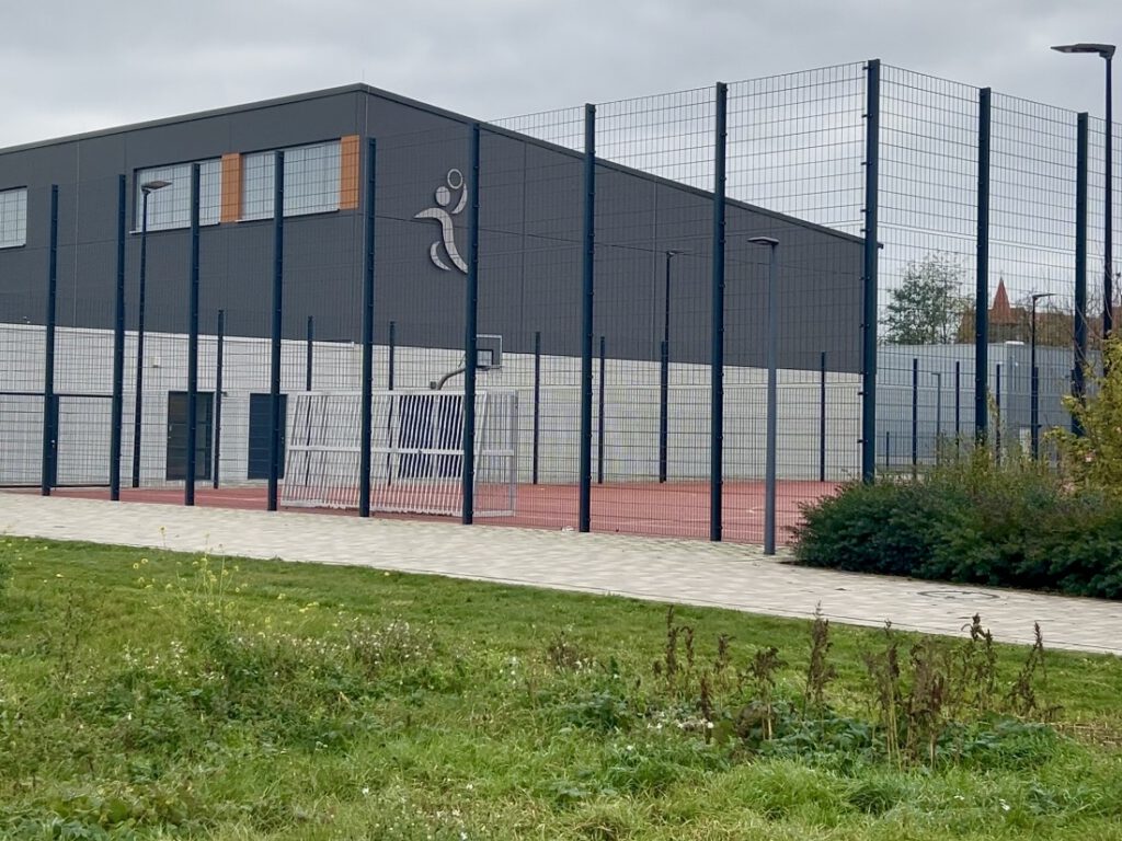 Blick von einer Grünfläche auf einen eingezäunten Bolzplatz vor einer Turnhalle